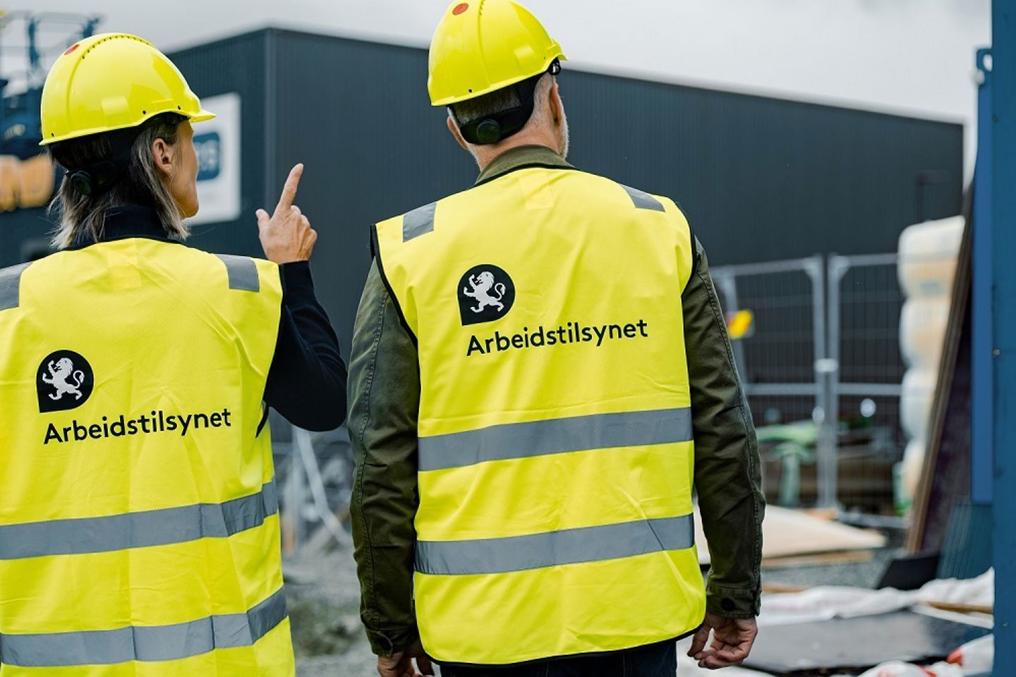 En kvinne og en mann på en byggeplass. Begge har på hjelm og vest med logo fra Arbeidstilsynet. Kvinnen peker på en byggeplass