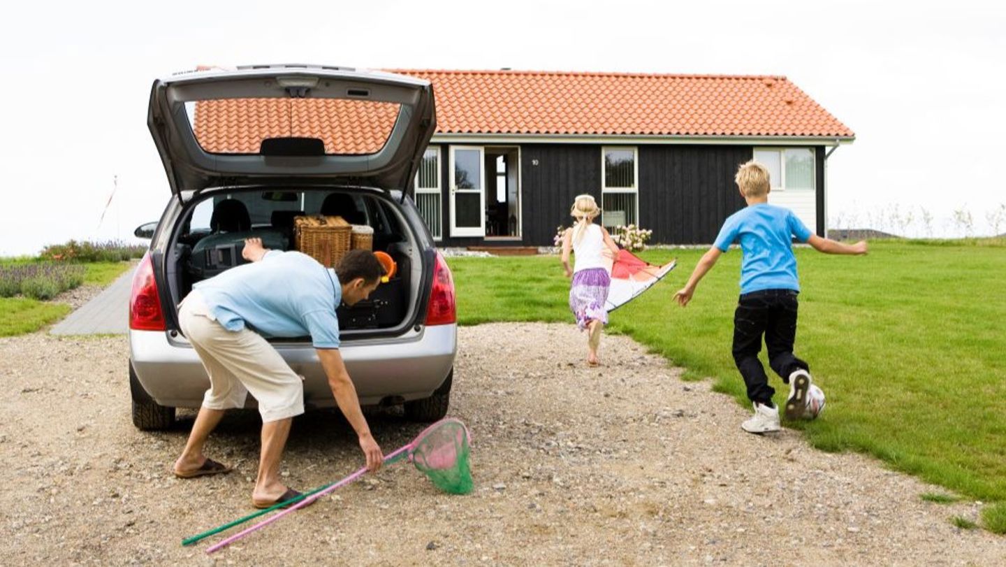Bildet viser en mann som pakker ut av en bil foran et feriehus. To barn løper fra bilen til huset. 