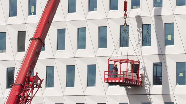 En arbeider henger i en kurv fra en kran utenfor et stort bygg